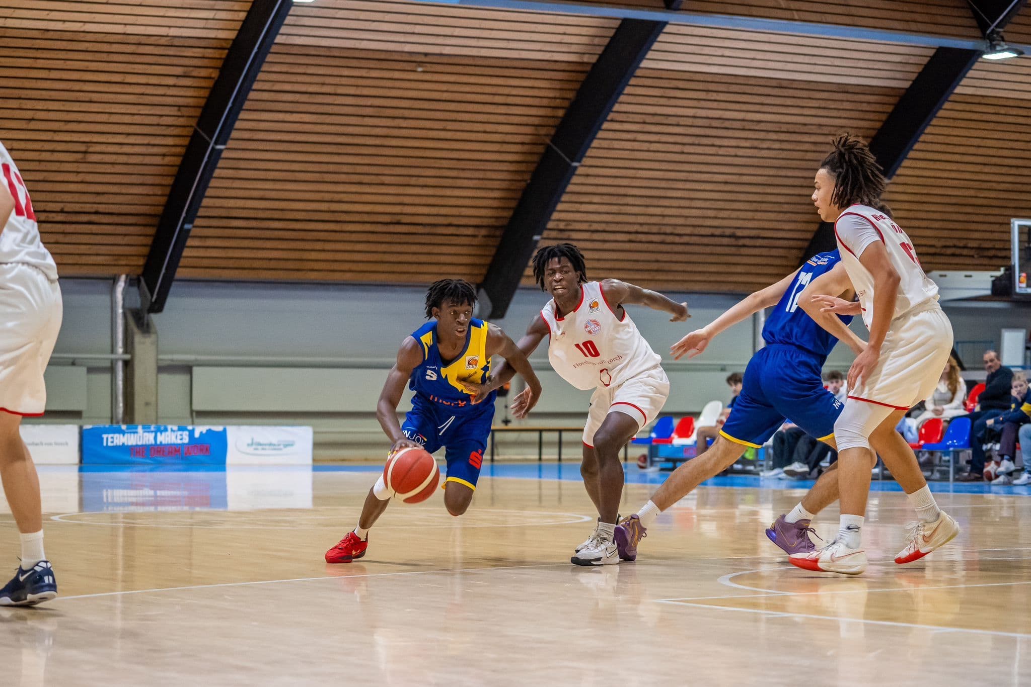 Ein Basketballspieler im blauen Trikot dribbelt, während andere Spieler folgen. Dynamische Szene.