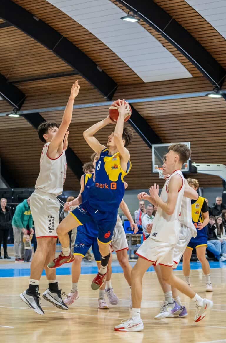 Ein Basketballspieler springt zum Wurf, umgeben von verteidigenden Gegenspielern in einer Sporthalle.