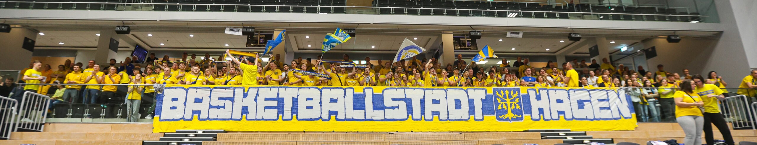 Fans in gelben T-Shirts jubeln begeistert in einer Basketballhalle, halten Schilder und Fahnen.