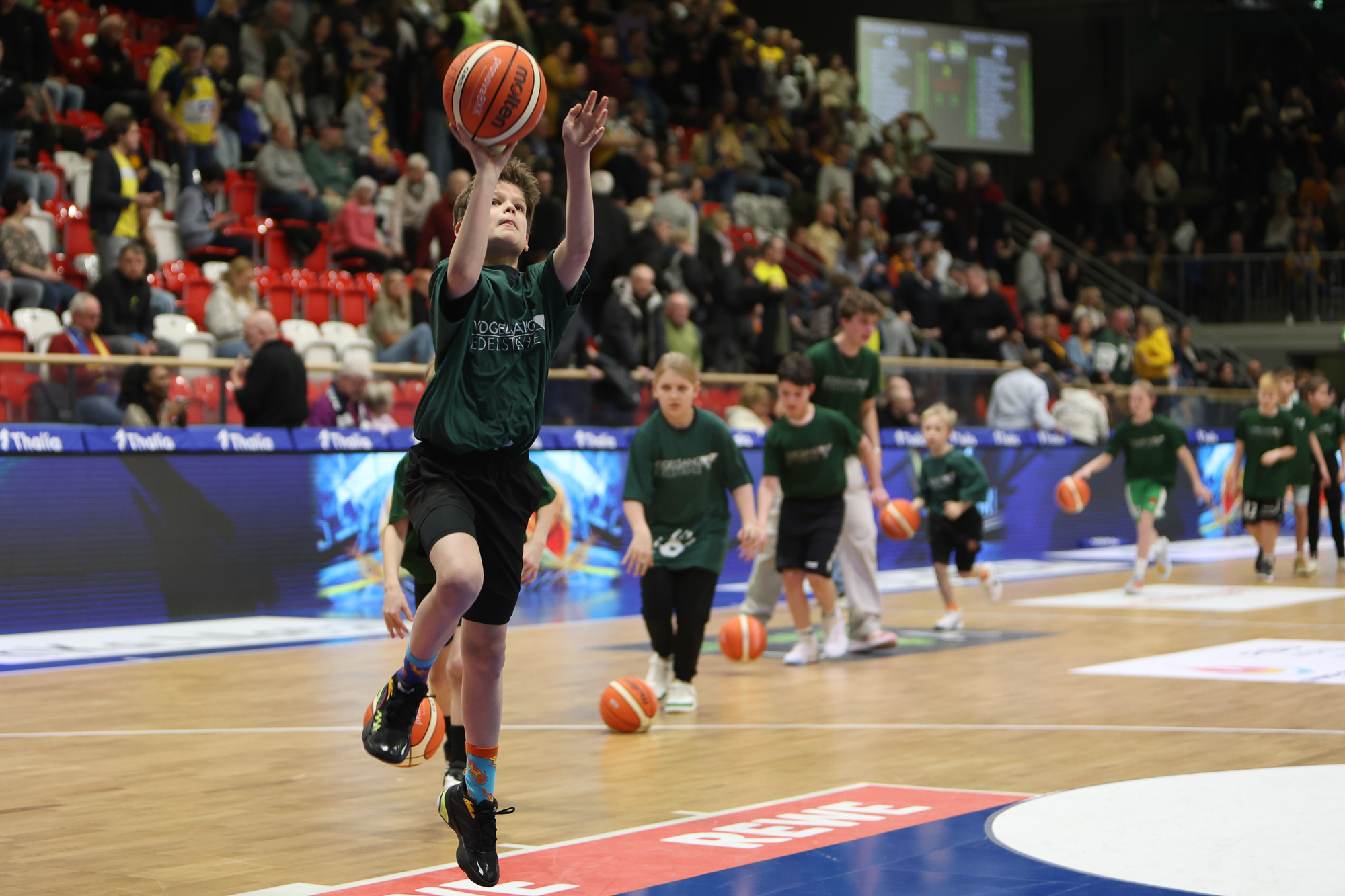 Junge beim Basketball-Wurf, energiegeladen, in grünem Shirt, bunte Socken, vor Publikum in einer Halle.