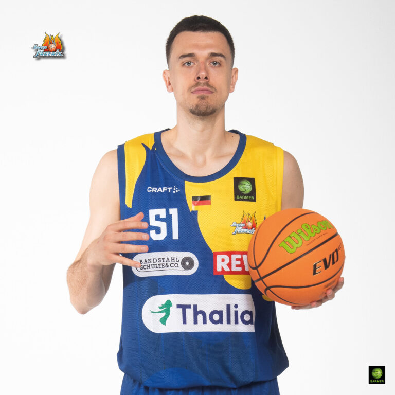 Ein Mann im blauen und gelben Basketballtrikot hält einen Basketball, schaut ernst.