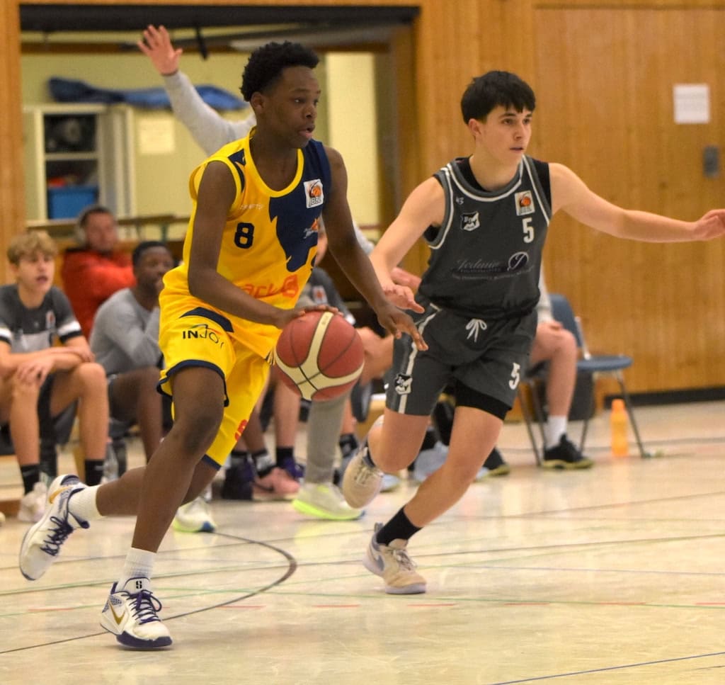 Basketballspieler im gelben Trikot dribbelt, verfolgt von Spieler im grauen Trikot in Sporthalle.