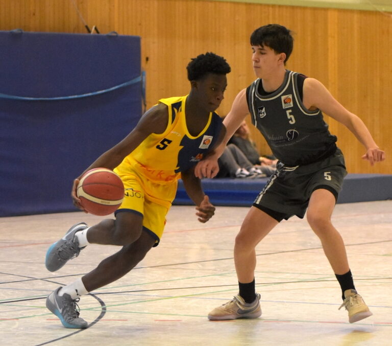 Zwei Basketballspieler in Aktion, einer in gelbem Trikot dribbelt, der andere in grauem verteidigt ihn.