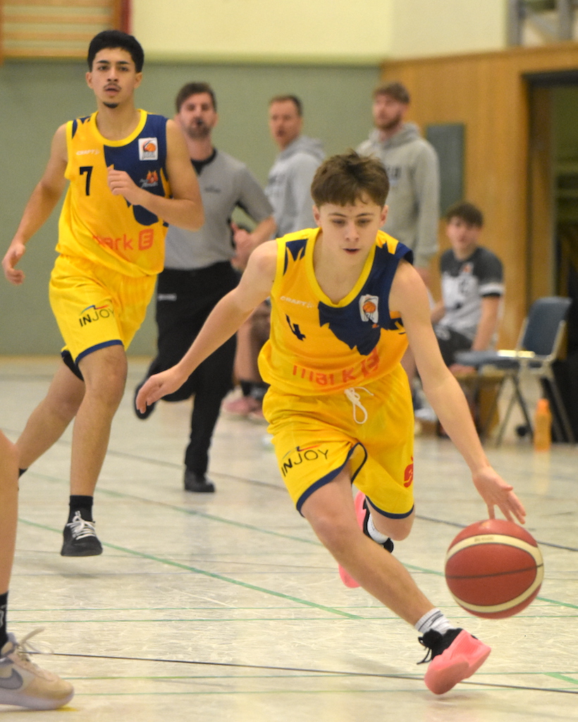 Junger Basketballspieler in gelbem Trikot dribbelt konzentriert, während andere Spieler zuschauen.