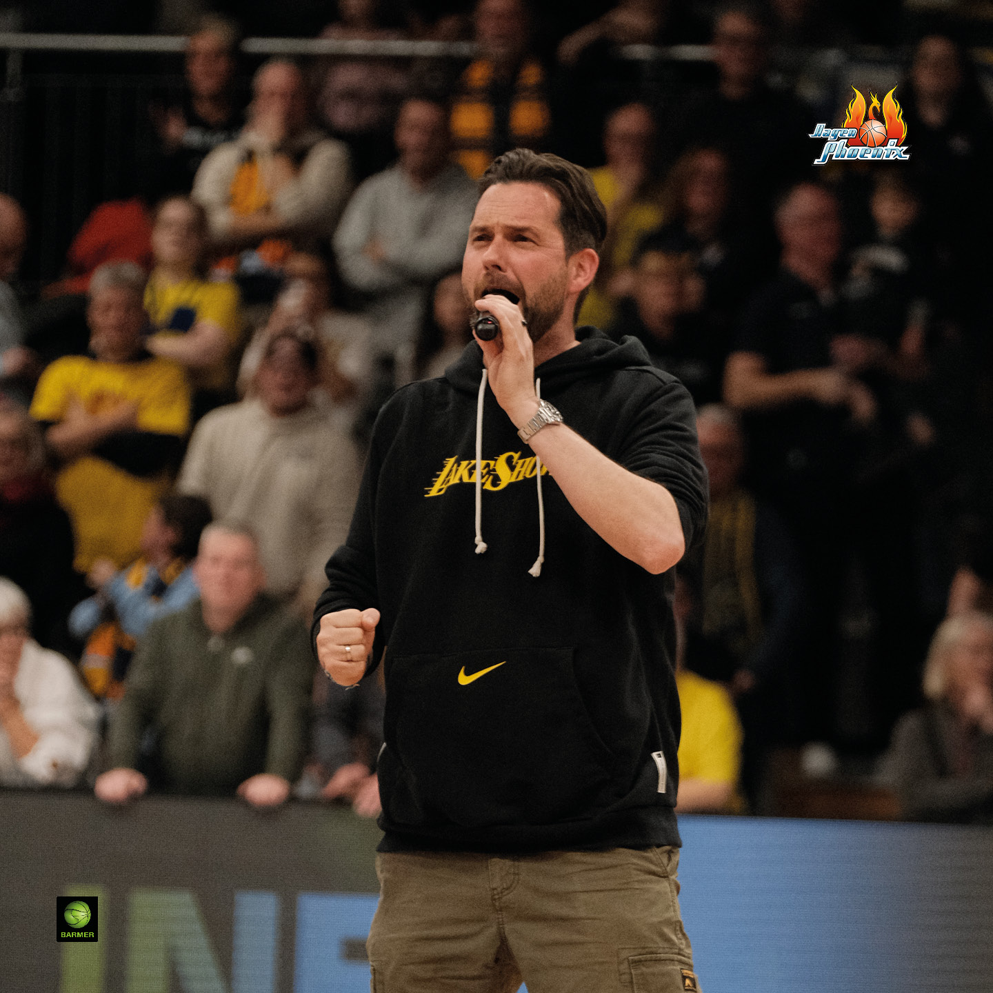 Ein Mann singt leidenschaftlich in einem belebten Indoor-Sportstadion, trägt schwarzen Hoodie.