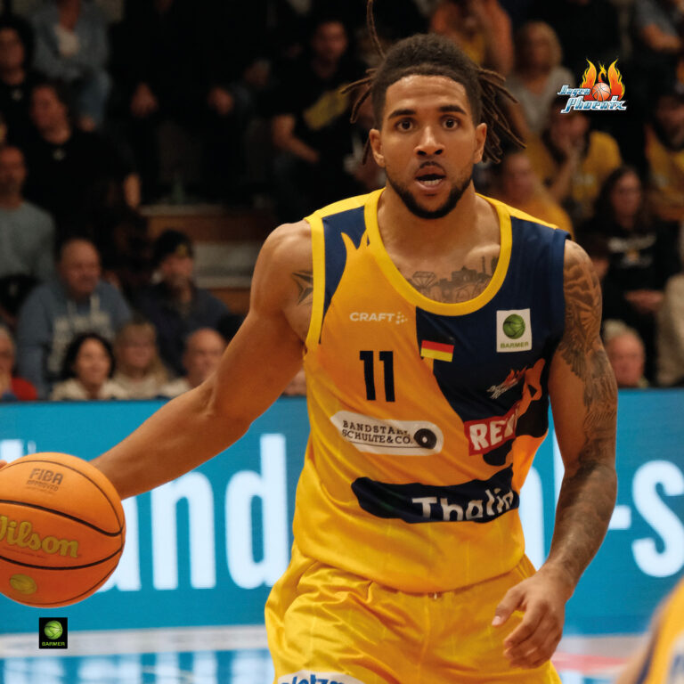 Ein Basketballspieler im gelben Trikot mit fokussiertem Ausdruck hält den Ball auf einem Spielfeld.