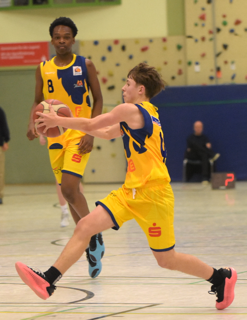 Ein Spieler in gelbem Trikot springt beim Basketball, ein weiterer schaut zu, helle Halle.