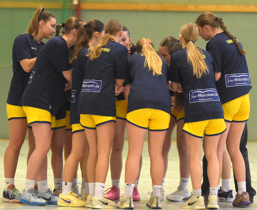 Handballteam in blauen Trikots und gelben Shorts, im Kreis stehend, fokussierte Ausdrücke.