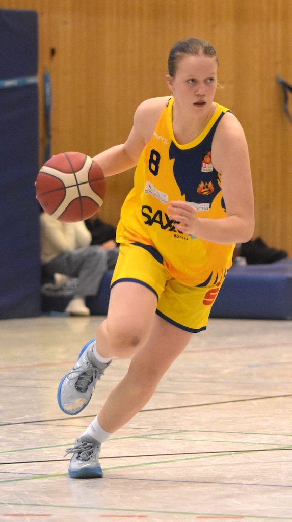 Basketballspielerin in gelbem Trikot dribbelt konzentriert den Ball auf einem Hallenparkett.