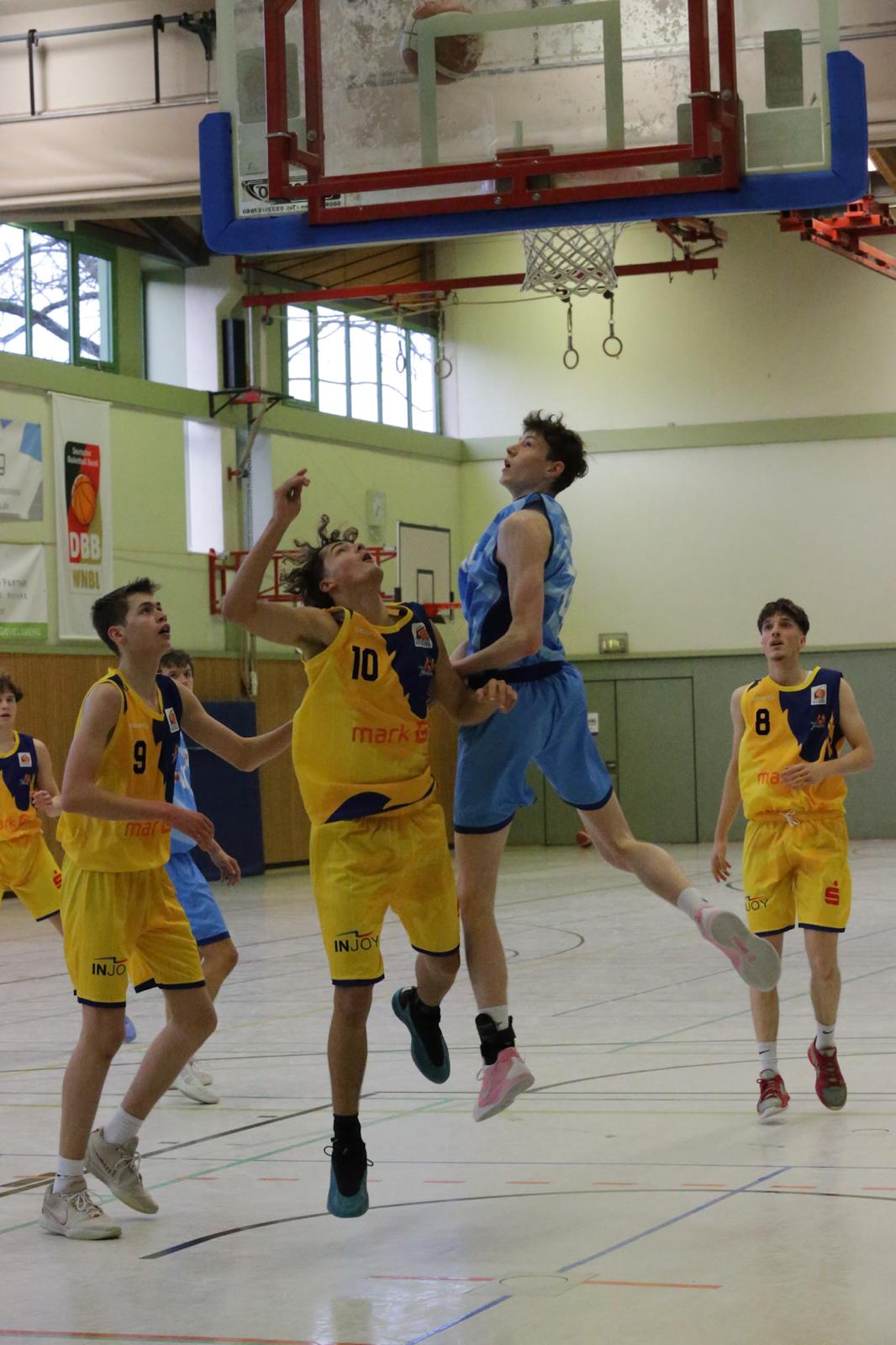 Basketballspiel mit Jugendlichen, spannender Moment, Spieler in blauen und gelben Trikots unter Korb.