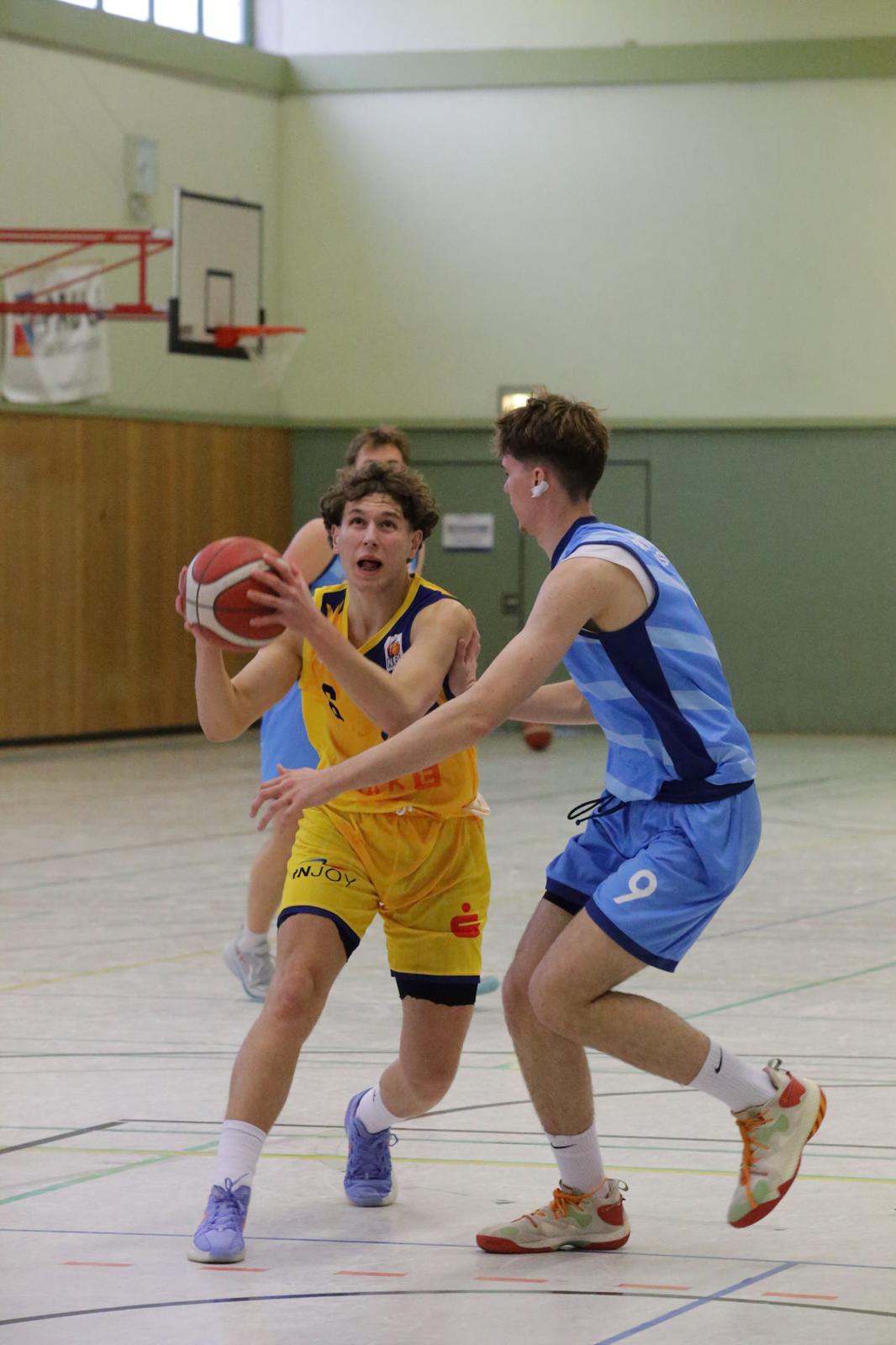 Zwei Basketballspieler, einer in gelb ernst, der andere in blau versucht ihn zu blocken, Hallenspiel.