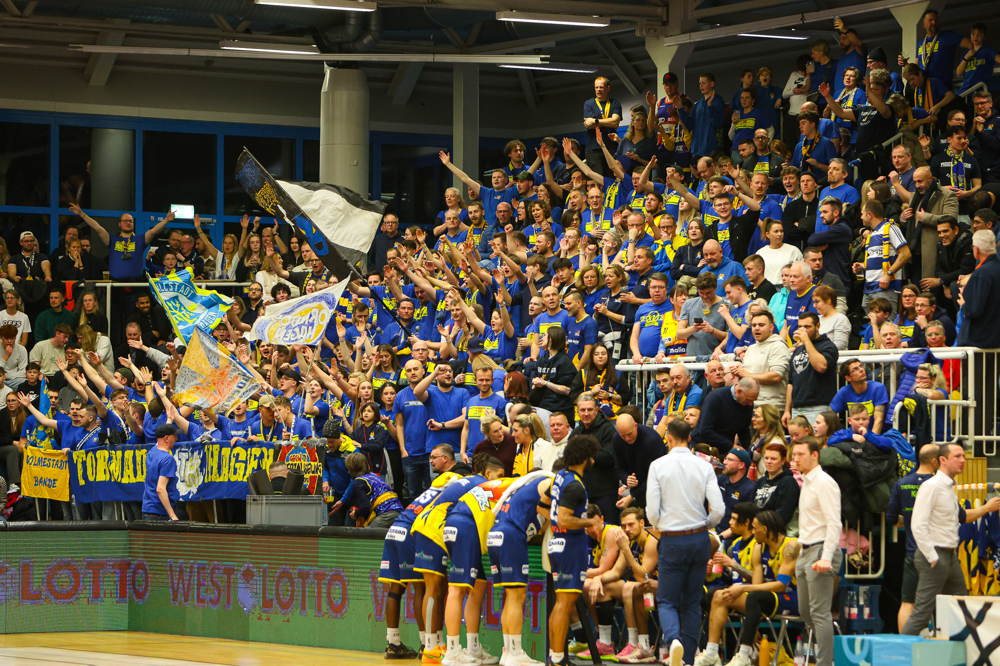 Basketballfans in blauer Kleidung jubeln im Stadion, während Spieler auf der Bank sitzen.