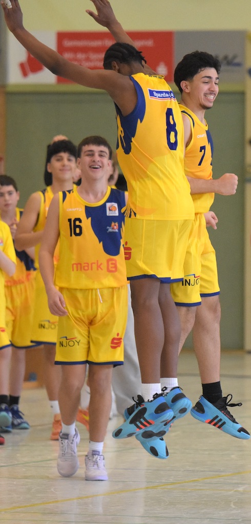 Zwei Basketballspieler in gelben Trikots jubeln, einer springt. Publikum im Hintergrund.