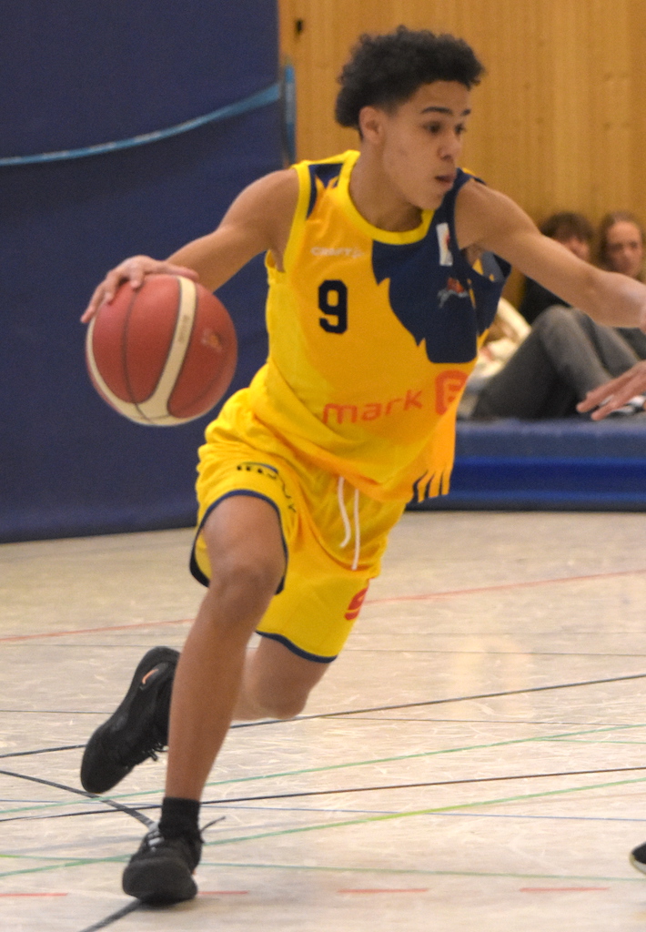 Basketballspieler in gelbem Trikot beim Dribbeln, fokussierter Ausdruck, Holzwand im Hintergrund.