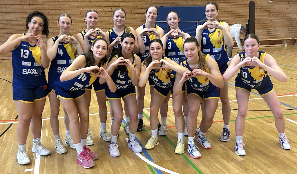 Eine fröhliche Mädchen-Basketballmannschaft posiert in blauen und gelben Trikots in einer Turnhalle.