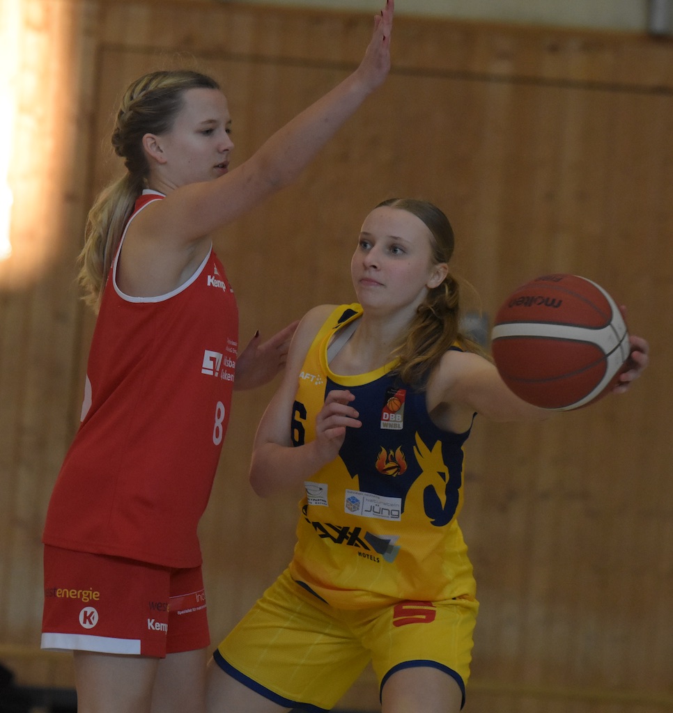 Zwei Frauen beim Basketballspiel, konzentriert, in roten und gelben Trikots, mit Holzwand im Hintergrund.