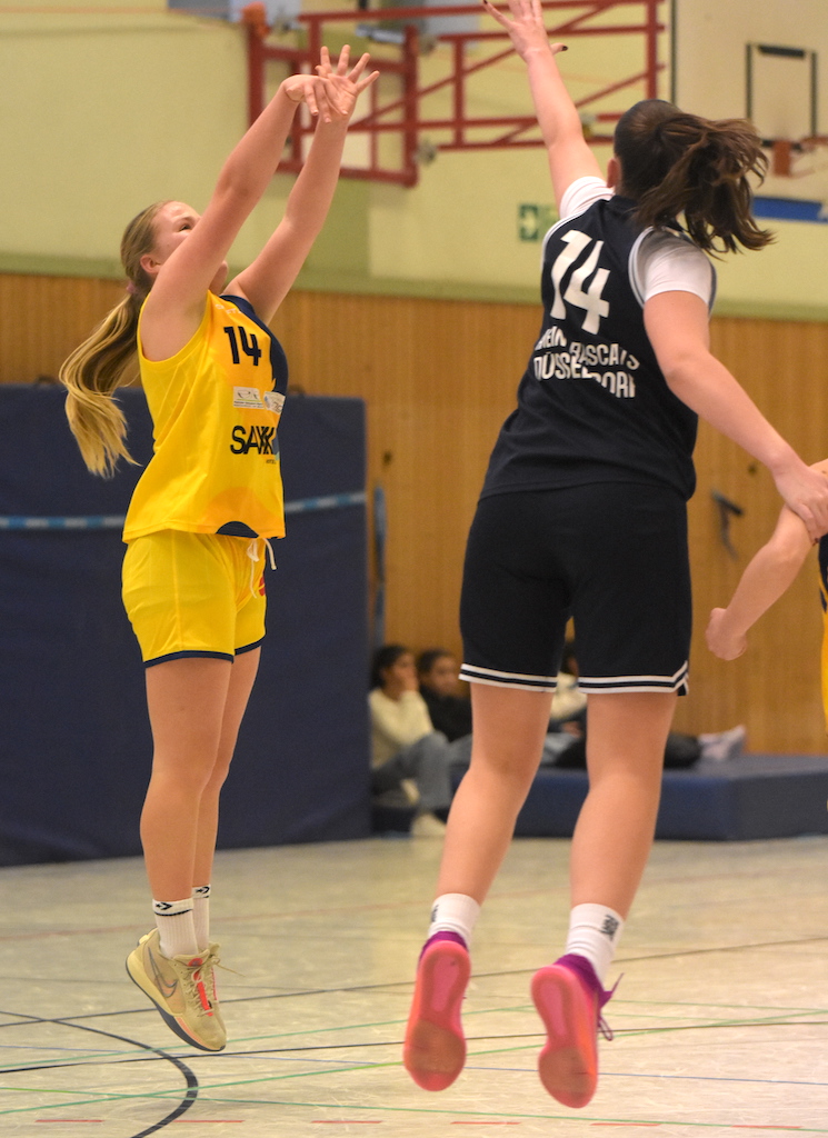 Zwei Basketballspielerinnen springen, eine in gelbem Trikot, die andere in schwarzem, in einer Halle.