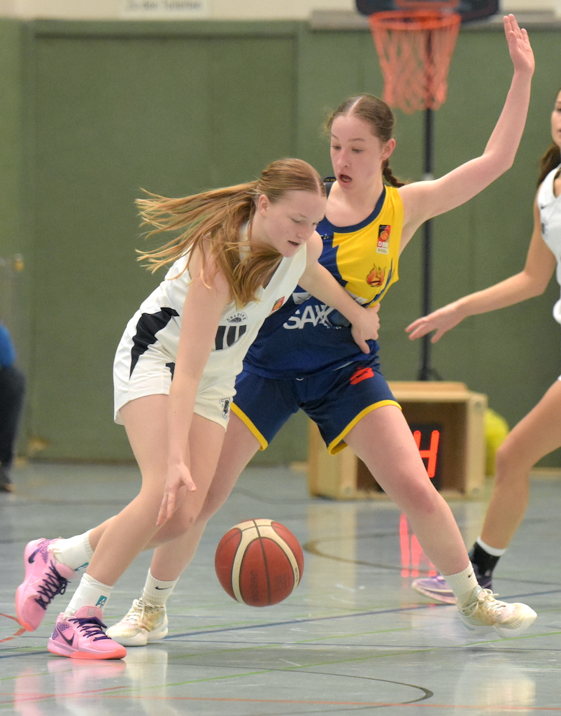 Zwei junge Basketballspielerinnen im Spiel, eine in weiß, eine in blau-gelb, mit konzentriertem Ausdruck.