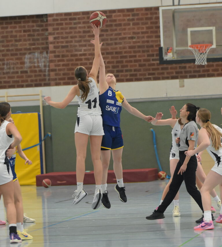 Basketballspiel; eine Spielerin in weiß springt mit entschlossenem Gesicht gegen eine in blau-gelb.