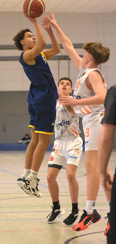 Ein junger Mann in blauem Trikot springt, um einen Basketball zu werfen. Zwei Verteidiger in weißen Trikots versuchen ihn zu blocken.