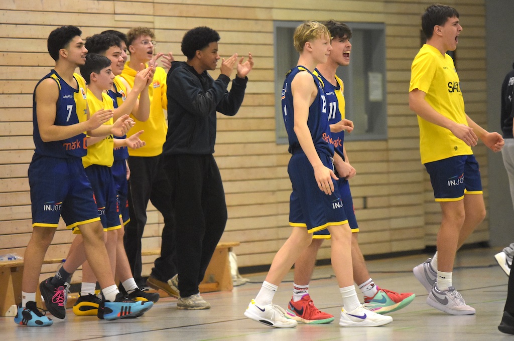 Basketballteam jubelt am Spielfeldrand; blaue Trikots, gelbe Akzente, Holzwand im Hintergrund.