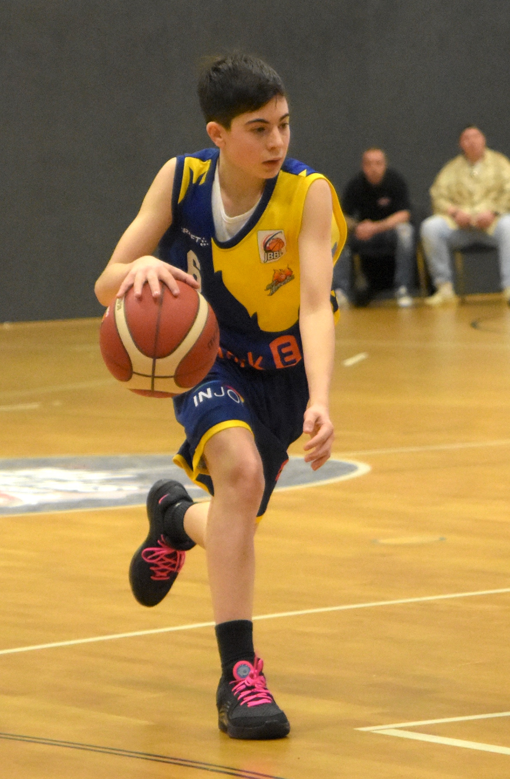 Junger Basketballspieler dribbelt konzentriert auf Holzboden in blau-gelbem Trikot.