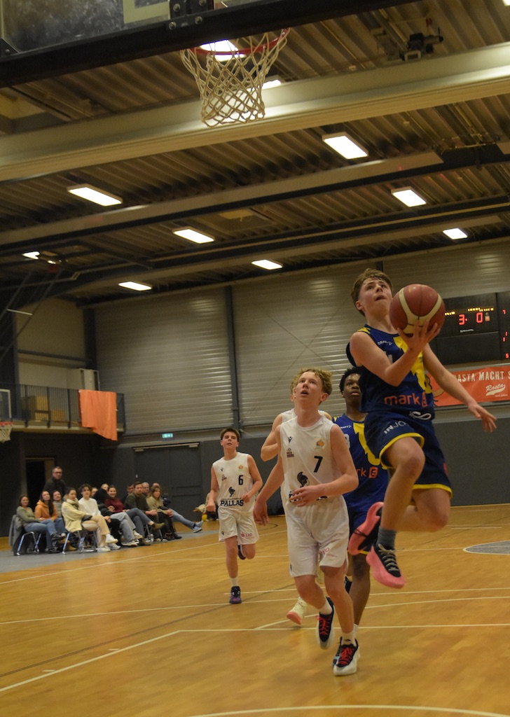 Basketballspiel, junger Spieler im blauen Trikot beim Korbleger. Zuschauer im Hintergrund.