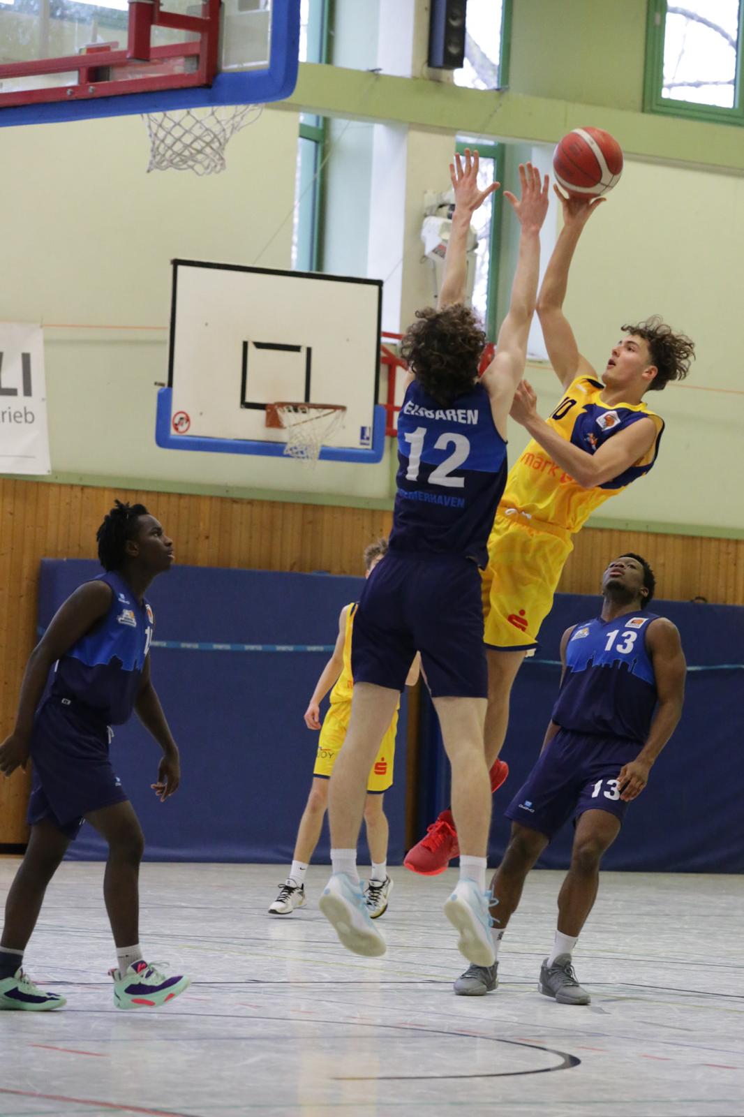 Ein Basketballspieler in gelbem Trikot springt zum Wurf über zwei Spieler in blauen Trikots.